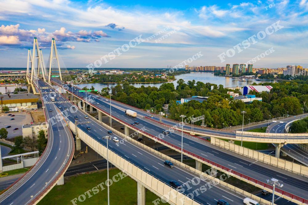 Фотография современного Санкт-Петербург, Россия, снятая с дрона. На изображении показано интенсивное движение транспорта: автомагистраль и дорожная развязка, проходящие по мосту через реку Нева. В кадре запечатлены Обуховский мост и вантовый мост, являющиеся частью современной транспортной инфраструктуры города. Фотоснимок передаёт масштаб мегаполиса, динамику автомобильного трафика и развитие дорожной архитектуры Петербурга. Это изображение подчёркивает роль мостов и автомагистралей в жизни крупного города. Иллюстрация может использоваться как фото, картинка или слайд, посвящённые дорогам и урбанистике российских городов.